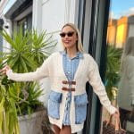 Dış mekânda beyaz triko hırka, kahverengi deri tokalı detaylar, mavi denim elbise ve kahverengi deri çizmelerle kombinlenmiş bir kadın modeli.