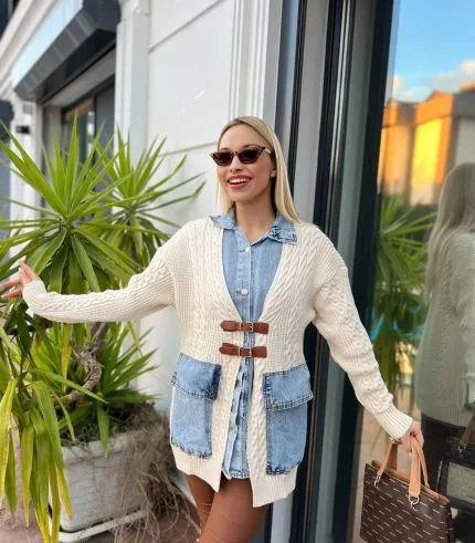 Dış mekânda beyaz triko hırka, kahverengi deri tokalı detaylar, mavi denim elbise ve kahverengi deri çizmelerle kombinlenmiş bir kadın modeli.