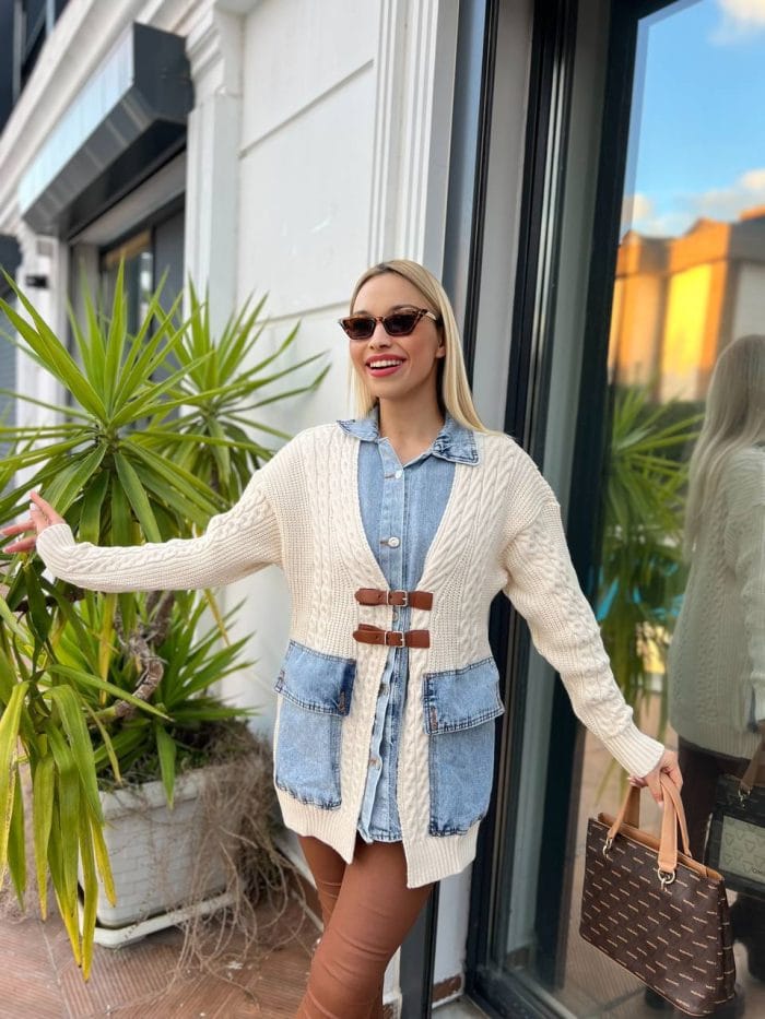 Dış mekânda beyaz triko hırka, kahverengi deri tokalı detaylar, mavi denim elbise ve kahverengi deri çizmelerle kombinlenmiş bir kadın modeli.