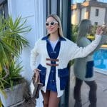 Dış mekânda beyaz triko hırka, kahverengi deri tokalı detaylar, mavi denim elbise ve kahverengi deri çizmelerle kombinlenmiş bir kadın modeli.