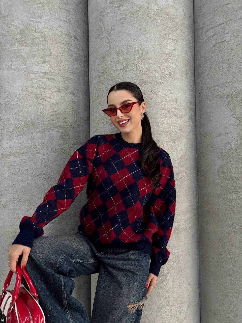 Ella Style lacivert baklava desenli oversize triko kazak giyen bir kadın. Kazağı, yırtık kot pantolon ve kırmızı detaylı bir çantayla kombinlemiş, kırmızı gözlüklerle şık bir dokunuş katmış.