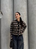 Ella Style pitikare ve baklava desenli siyah kadın hırka giyen bir kadın, bol kesim kot pantolon ve siyah çantayla kombin yapmış. Modern bir görünüm sağlayan gri beton sütunlar arka planda yer alıyor.