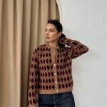 Ella Style pitikare ve baklava desenli siyah kadın hırka giyen bir kadın, bol kesim kot pantolon ve siyah çantayla kombin yapmış. Modern bir görünüm sağlayan gri beton sütunlar arka planda yer alıyor.