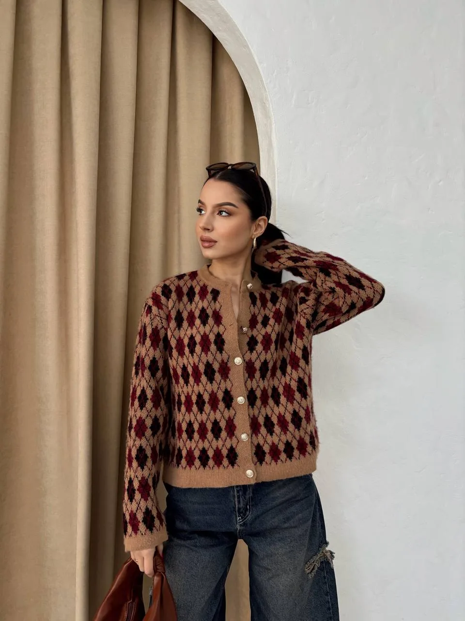 Ella Style pitikare ve baklava desenli siyah kadın hırka giyen bir kadın, bol kesim kot pantolon ve siyah çantayla kombin yapmış. Modern bir görünüm sağlayan gri beton sütunlar arka planda yer alıyor.