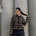 Ella Style pitikare ve baklava desenli siyah kadın hırka giyen bir kadın, bol kesim kot pantolon ve siyah çantayla kombin yapmış. Modern bir görünüm sağlayan gri beton sütunlar arka planda yer alıyor.