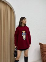 Ayıcık işlemeli bordo oversize kazak giyen bir kadın. Siyah mini etek, siyah çizmeler ve siyah el çantası ile kombinlenmiş.