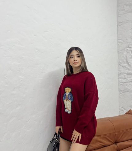 Ayıcık işlemeli bordo oversize kazak giyen bir kadın. Siyah mini etek, siyah çizmeler ve siyah el çantası ile kombinlenmiş.