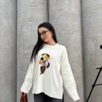 Seyahat eden ayıcık işlemeli beyaz kazak giyen bir kadın. Siyah pantolon, gözlük ve kahverengi çanta ile kombinlenmiş.