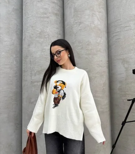 Seyahat eden ayıcık işlemeli beyaz kazak giyen bir kadın. Siyah pantolon, gözlük ve kahverengi çanta ile kombinlenmiş.