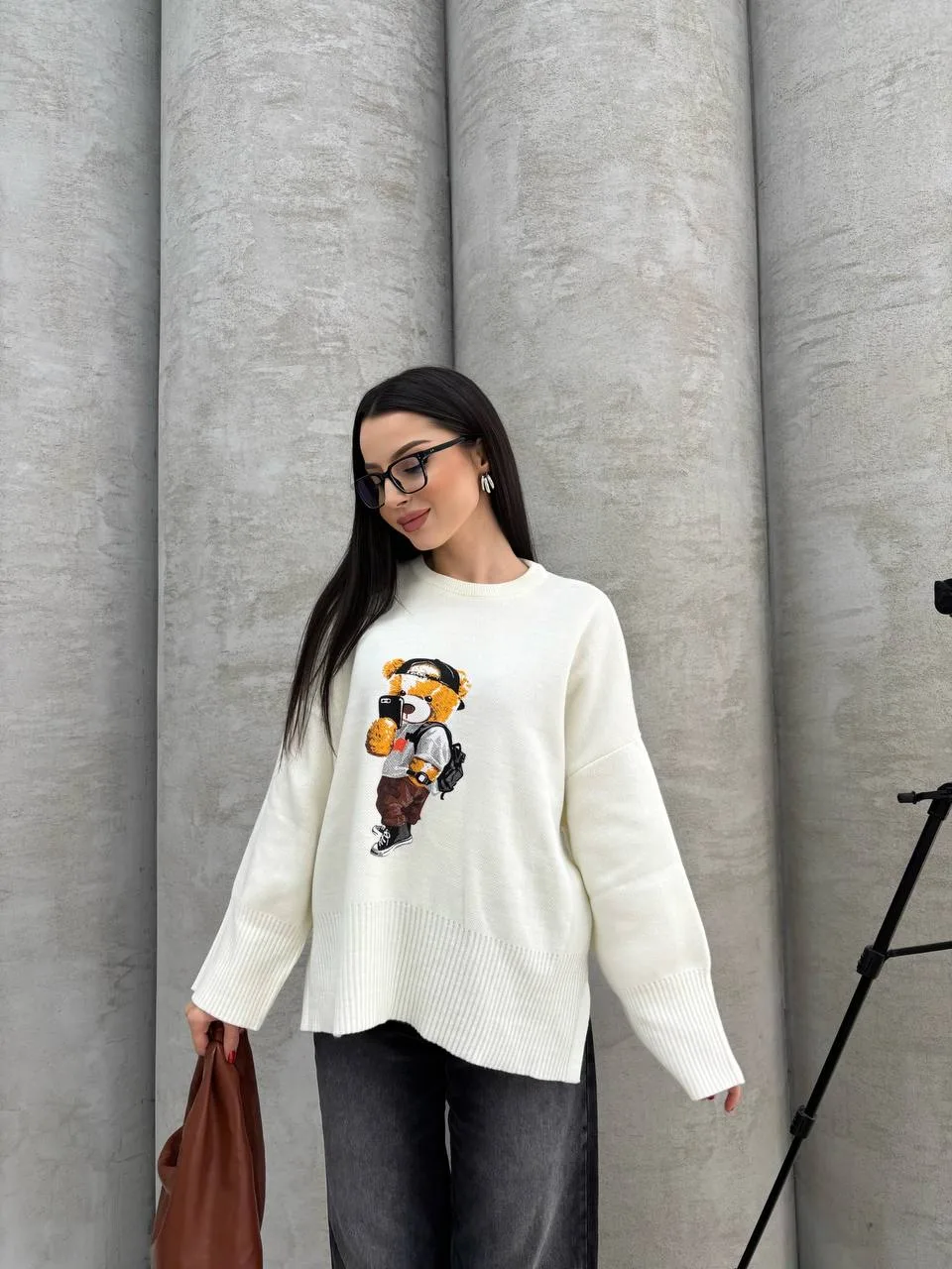 Seyahat eden ayıcık işlemeli beyaz kazak giyen bir kadın. Siyah pantolon, gözlük ve kahverengi çanta ile kombinlenmiş.