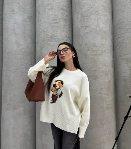 Seyahat eden ayıcık işlemeli beyaz kazak giyen bir kadın. Siyah pantolon, gözlük ve kahverengi çanta ile kombinlenmiş.