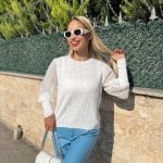 Şifon kollu örgü detaylı beyaz triko kazak giyen kadın, mavi deri görünümlü pantolon ve beyaz çantayla poz veriyor. Arka planda yeşilliklerle çevrili bahçe ve taş duvar bulunuyor.