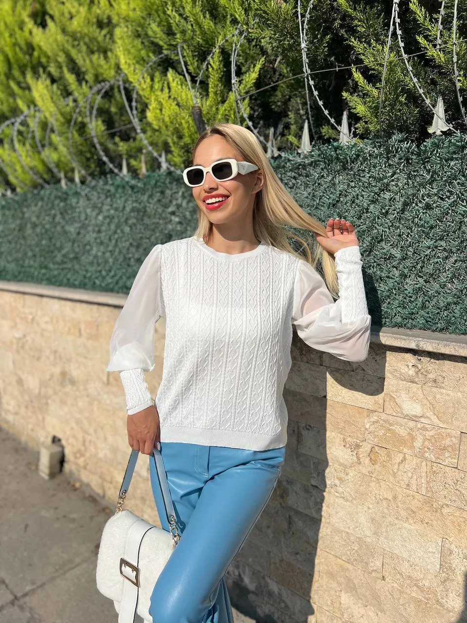 Şifon kollu örgü detaylı beyaz triko kazak giyen kadın, mavi deri görünümlü pantolon ve beyaz çantayla poz veriyor. Arka planda yeşilliklerle çevrili bahçe ve taş duvar bulunuyor.
