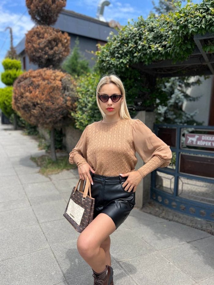 Şifon kollu örgü detaylı beyaz triko kazak giyen kadın, mavi deri görünümlü pantolon ve beyaz çantayla poz veriyor. Arka planda yeşilliklerle çevrili bahçe ve taş duvar bulunuyor.