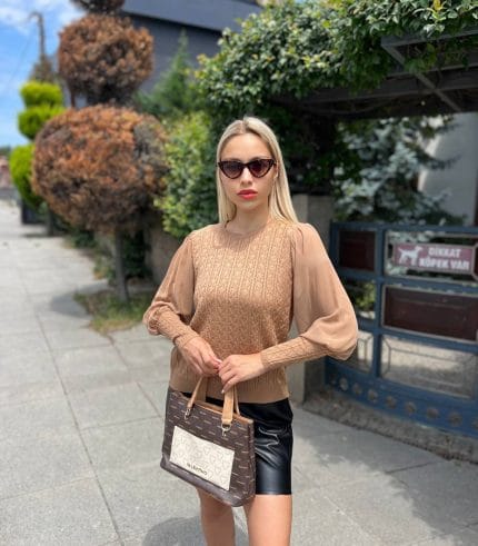 Şifon kollu örgü detaylı beyaz triko kazak giyen kadın, mavi deri görünümlü pantolon ve beyaz çantayla poz veriyor. Arka planda yeşilliklerle çevrili bahçe ve taş duvar bulunuyor.