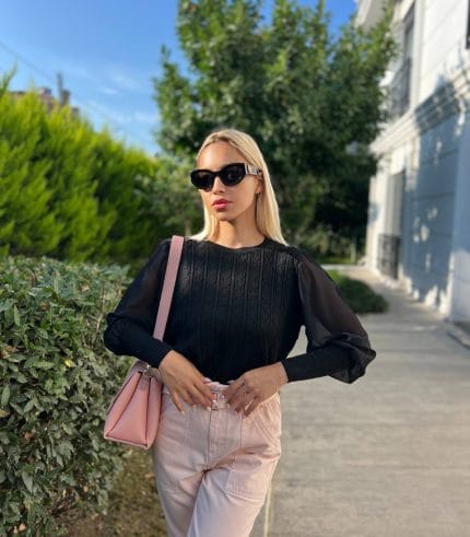 Şifon kollu örgü detaylı beyaz triko kazak giyen kadın, mavi deri görünümlü pantolon ve beyaz çantayla poz veriyor. Arka planda yeşilliklerle çevrili bahçe ve taş duvar bulunuyor.