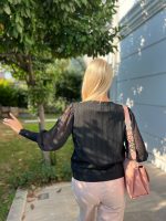 Şifon kollu örgü detaylı beyaz triko kazak giyen kadın, mavi deri görünümlü pantolon ve beyaz çantayla poz veriyor. Arka planda yeşilliklerle çevrili bahçe ve taş duvar bulunuyor.