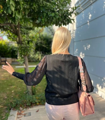 Şifon kollu örgü detaylı beyaz triko kazak giyen kadın, mavi deri görünümlü pantolon ve beyaz çantayla poz veriyor. Arka planda yeşilliklerle çevrili bahçe ve taş duvar bulunuyor.