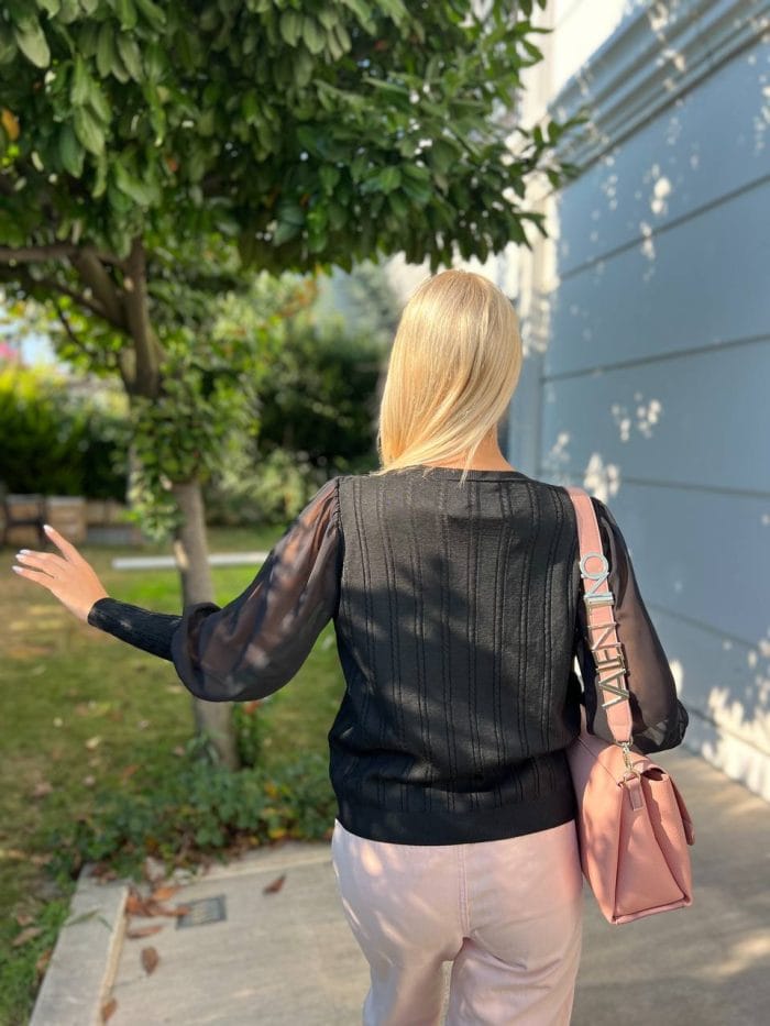 Şifon kollu örgü detaylı beyaz triko kazak giyen kadın, mavi deri görünümlü pantolon ve beyaz çantayla poz veriyor. Arka planda yeşilliklerle çevrili bahçe ve taş duvar bulunuyor.