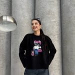 Siyah renkte, üzerinde ayıcık işlemeli detay bulunan oversize kazak giyen bir kadın. Yırtık mavi kot pantolon ve gözlükle kombinlenmiş.