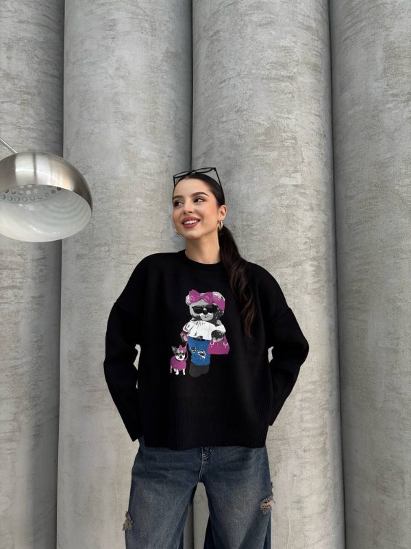 Siyah renkte, üzerinde ayıcık işlemeli detay bulunan oversize kazak giyen bir kadın. Yırtık mavi kot pantolon ve gözlükle kombinlenmiş.