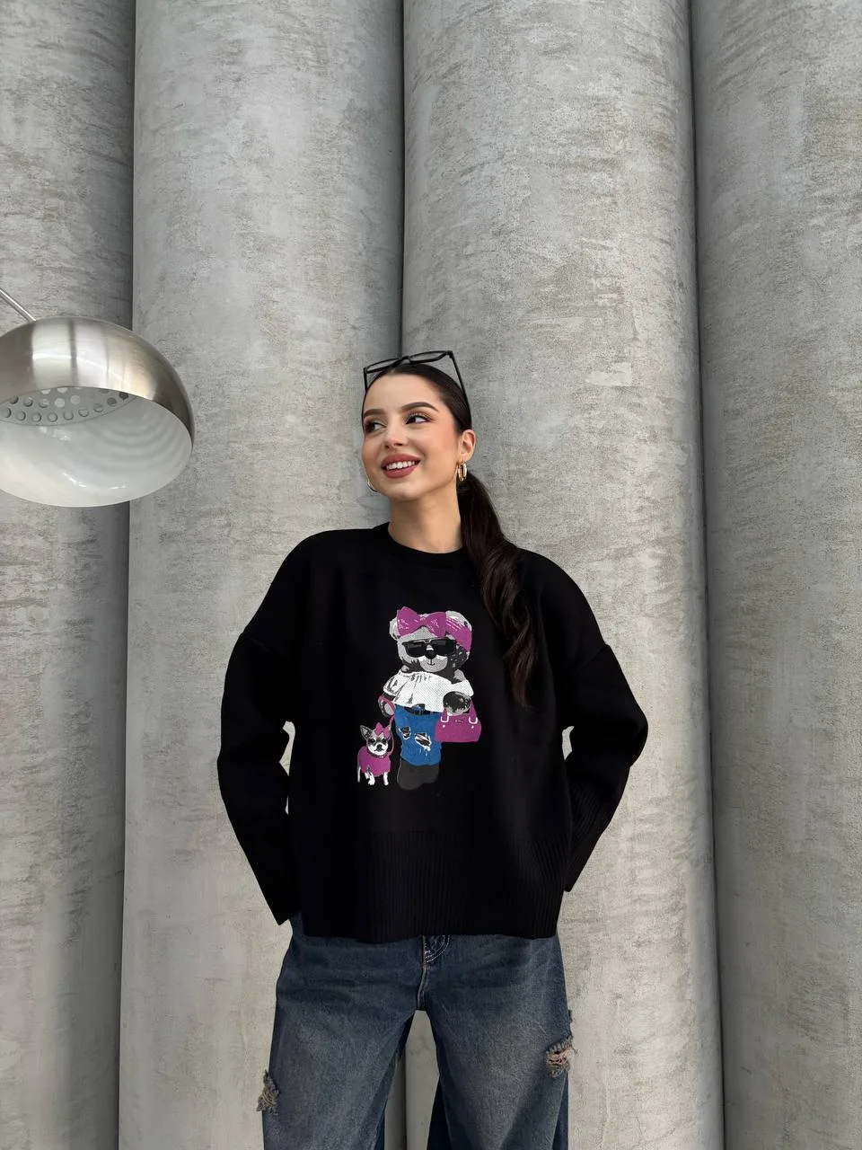 Siyah renkte, üzerinde ayıcık işlemeli detay bulunan oversize kazak giyen bir kadın. Yırtık mavi kot pantolon ve gözlükle kombinlenmiş.