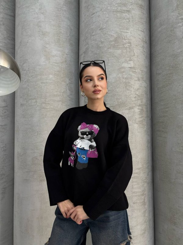 Siyah renkte, üzerinde ayıcık işlemeli detay bulunan oversize kazak giyen bir kadın. Yırtık mavi kot pantolon ve gözlükle kombinlenmiş.