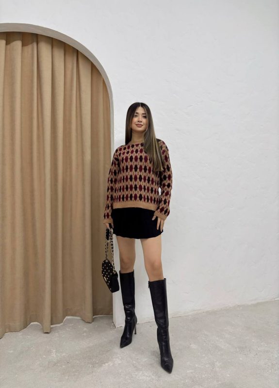 Baklava desenli bordo ve bej tonlarında yün kazak giyen bir kadın. Siyah mini etek, siyah çizme ve zincir detaylı siyah çantayla kombinlenmiş.