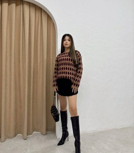 Baklava desenli bordo ve bej tonlarında yün kazak giyen bir kadın. Siyah mini etek, siyah çizme ve zincir detaylı siyah çantayla kombinlenmiş.