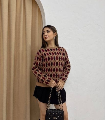 Baklava desenli bordo ve bej tonlarında yün kazak giyen bir kadın. Siyah mini etek, siyah çizme ve zincir detaylı siyah çantayla kombinlenmiş.