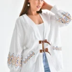 Beyaz zemin üzerinde, nakış detaylı tokalı hırka giyen kadın. Şık bir stil oluşturan zarif nakışlar ve modern kesim.