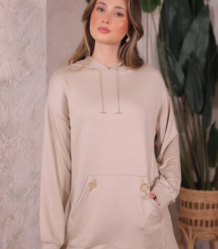 Doğal tonlarda tasarlanmış iç mekânda Oysho kumaş ikili takım giyen kadın. Kapüşonlu üst ve rahat kesim pantolonla şık bir kombin.